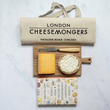 A small selection of cheese on a wooden board, with a cheese knife, book about cheese and a tote bag.