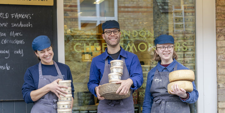 Three cheesemongers in their uniforms holding cheeses and laughing together outside a cheese shop.