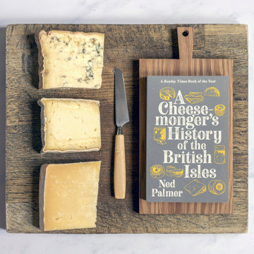 Three cuts of cheese on a wooden board, with a cheese knife and a paperback copy of Ned Palmer's 'A Cheesemonger's History of the British Isles'.
