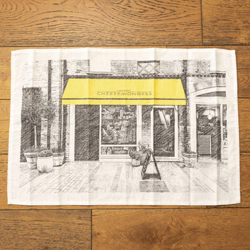 A black and white drawing of a cheese shop printed on a tea towel, placed on a wooden floor.