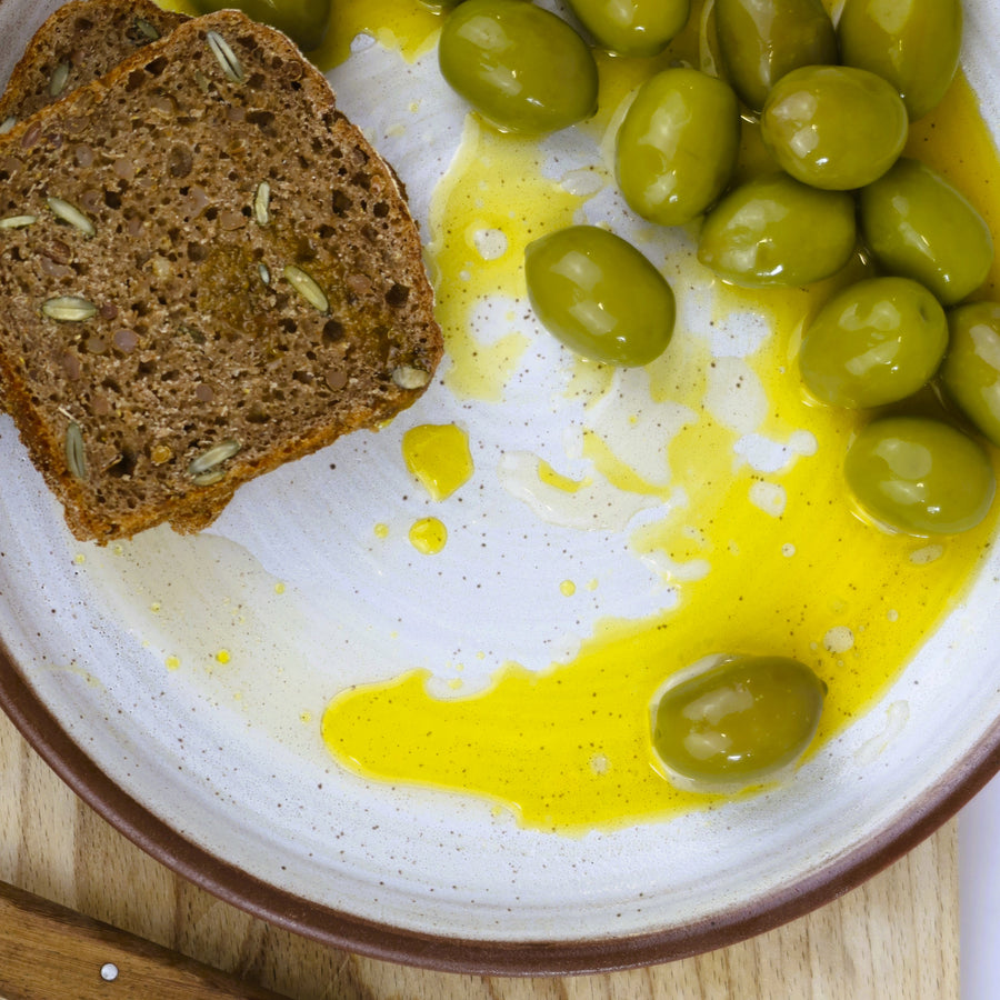 Overhead shot of Intosso green olives on a plate, with a slice of rye bread on the side and drizzled with some green extra virgin olive oil.