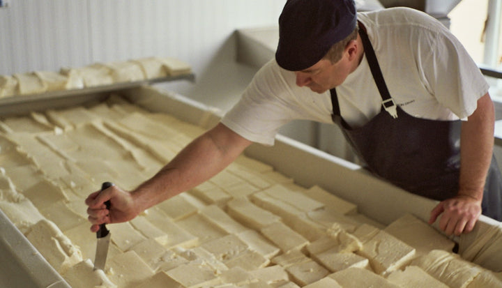A man, dressed in a white t-shirt, apron and cap, cutting the curds in the making of Stichelton blue cheese.