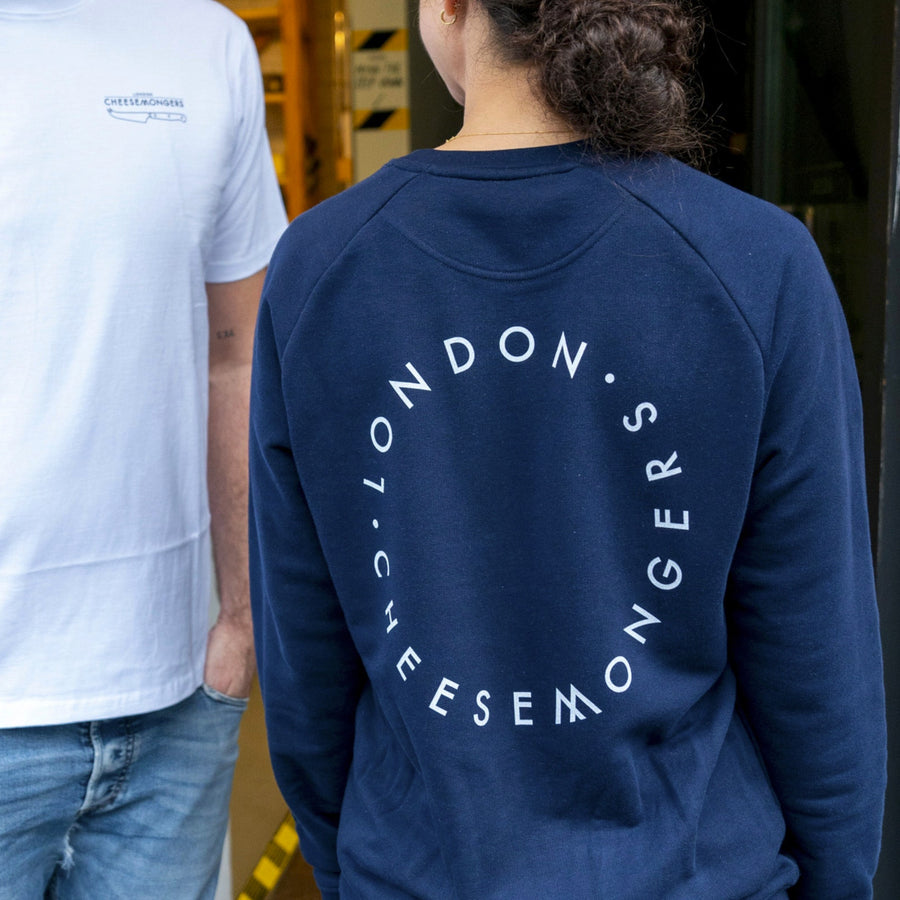 The back of a lady wearing a navy sweatshirt, with a round London Cheesemongers logo on the back, standing next to a man wearing a white t-shirt.