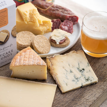 A selection of cut cheeses and a glass of beer on a wooden board.