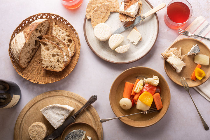 A table laid out with plates of cheese and pickles, a basket of bread and a board of several other cheeses