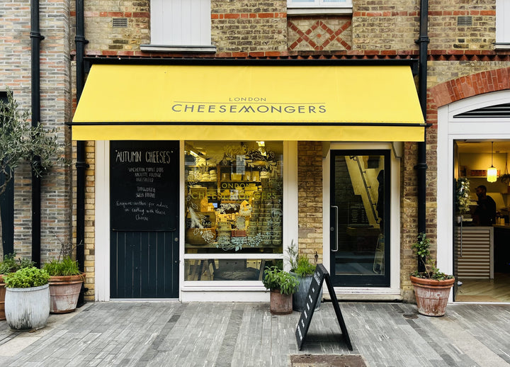 The front of a cheese shop with a yellow branded awning on a pedestrian street.