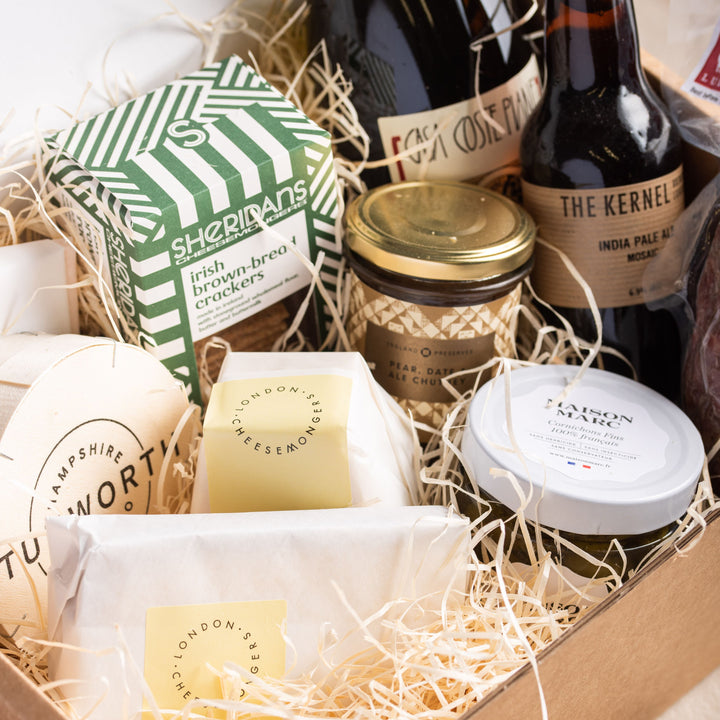 A selection of wrapped pieces of cheese and jars of accompaniments packedc with wood wool in a cardboard box.