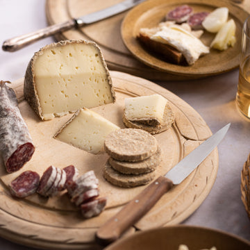 A selection of cuts of cheese with some slices of salami and oat biscuits with a knife on a wooden board.