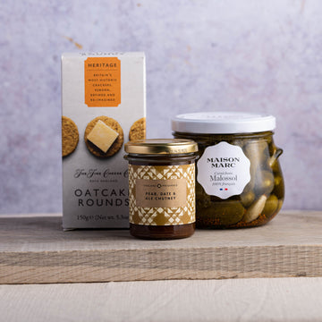 A packet of biscuits for cheese, a jar of cornichons and a jar of chutney on a wooden board.