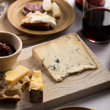A slice of Stilton blue cheese on a wooden board next to some Cheddar cheese on a slice of bread, with plates of accompaniments.