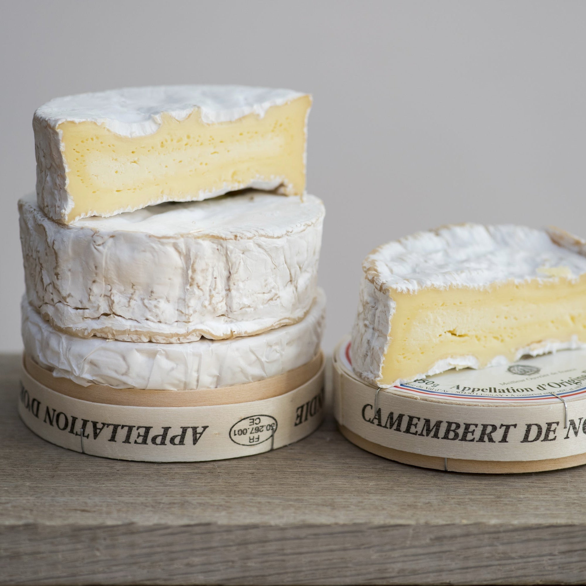 Three wheels of Camembert de Normandie cheese on a wooden board with a grey background.