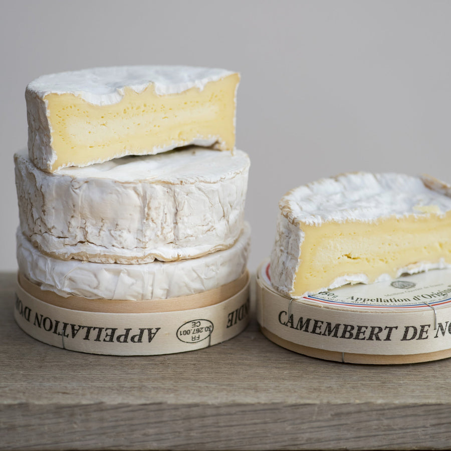 Three wheels of Camembert de Normandie cheese on a wooden board with a grey background.