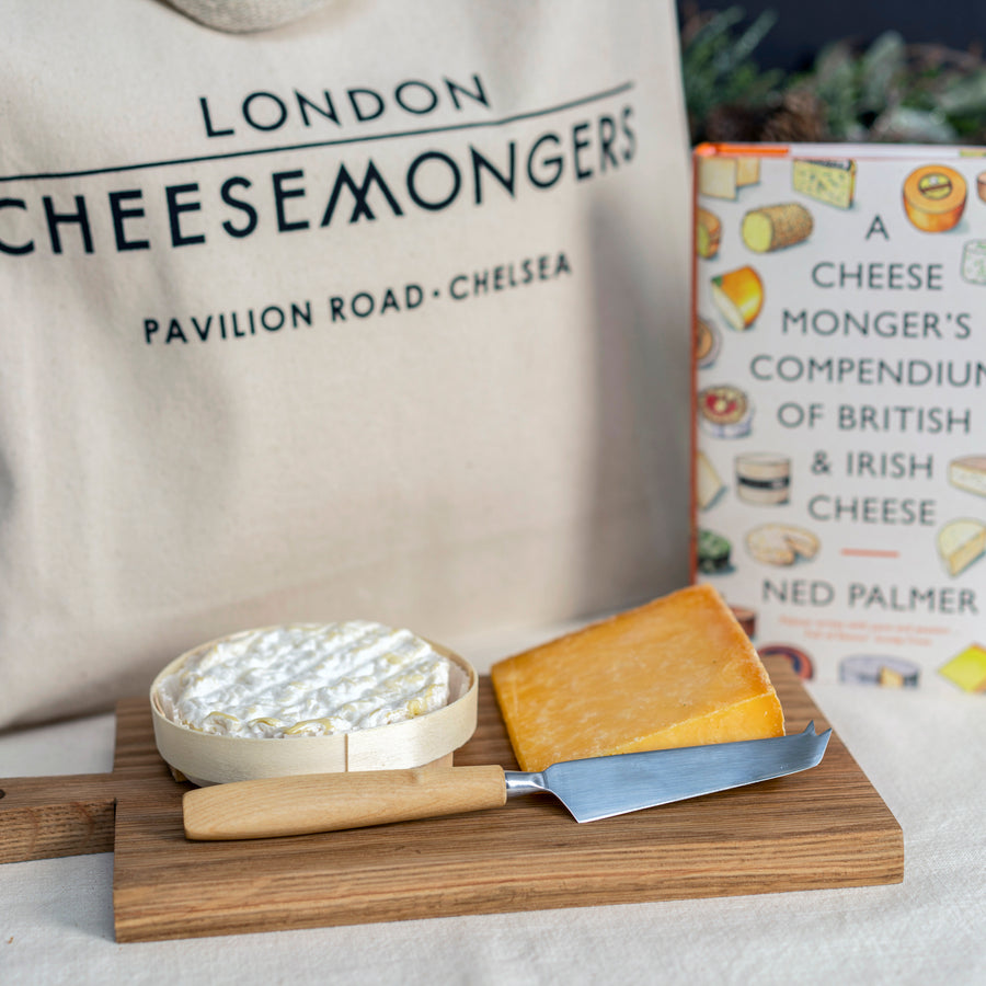 A whole St. Felicien, a cut of Cheshire cheese with a cheese knife on a small wooden board on a table in front of a tote bag and a book about cheese. 