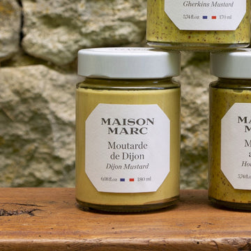 Glass jars of French mustards on a wooden table in frront of a stone wall.