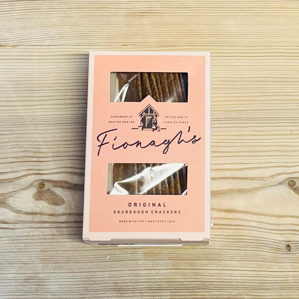 A package of Fionagh's Original Sourdough Crackers placed on a light wood grain surface. The packaging is a soft peach-pink color with a clear window showing the brown crackers inside, and the brand name 'Fionagh's' is written in black script.