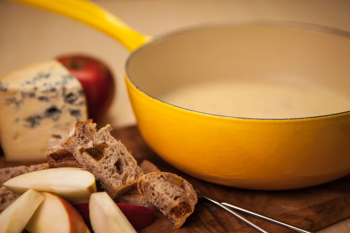 Cut pieces of fruit, bread and cheese on a wooden board with a pan of warm fondue. 