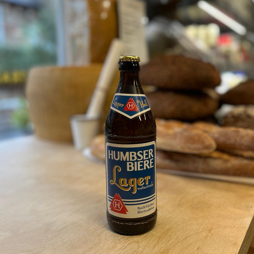A bottle of Humbser Biere Lager beer standing on a light wooden counter in a bakery setting, with bread loaves blurred in the background.