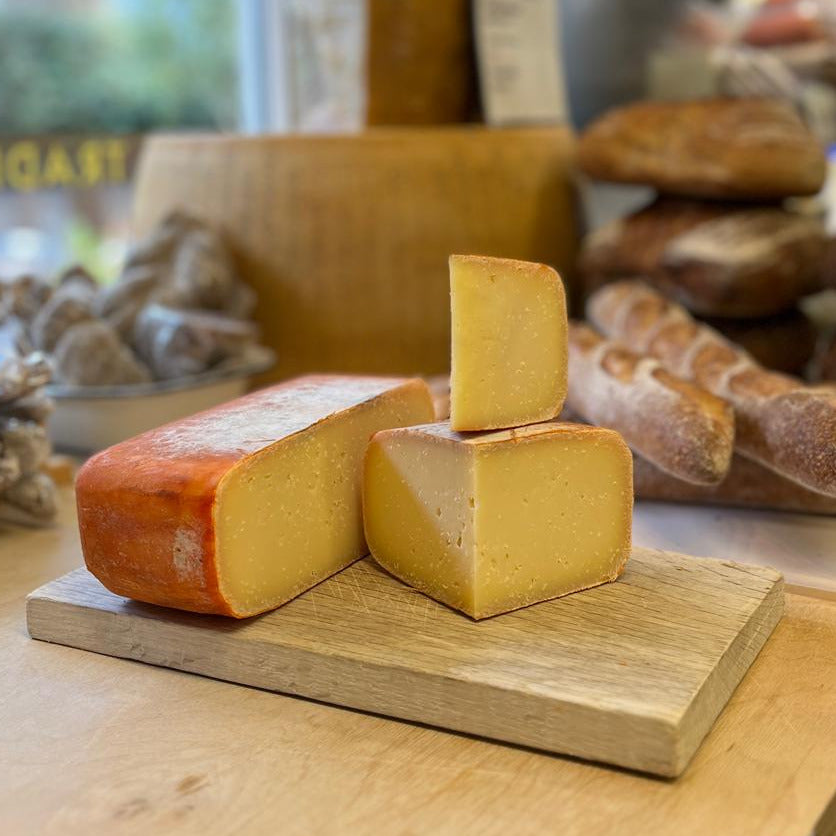 A close-up of artisanal cheeses on a wooden board in a bakery or cheese shop, with rustic bread and shelves blurred in the background.
