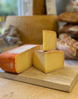 A close-up of artisanal cheeses on a wooden board in a bakery or cheese shop, with rustic bread and shelves blurred in the background.