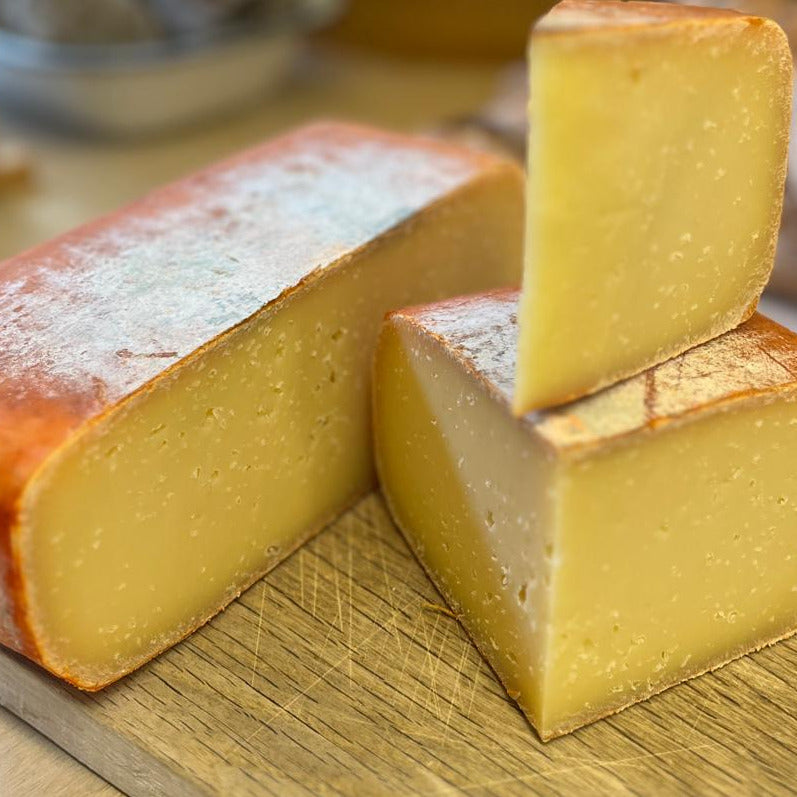 Close-up of a rustic wedge and slice of golden yellow cheese with a reddish-brown rind, resting on a wooden cutting board.