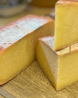 Close-up of a rustic wedge and slice of golden yellow cheese with a reddish-brown rind, resting on a wooden cutting board.