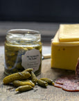 A jar of Maison Marc Extra Fins cornichons on a wooden board with some sliced salami and cheese.