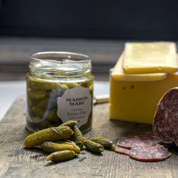 A jar of Maison Marc Extra Fins cornichons on a wooden board with some sliced salami and cheese.