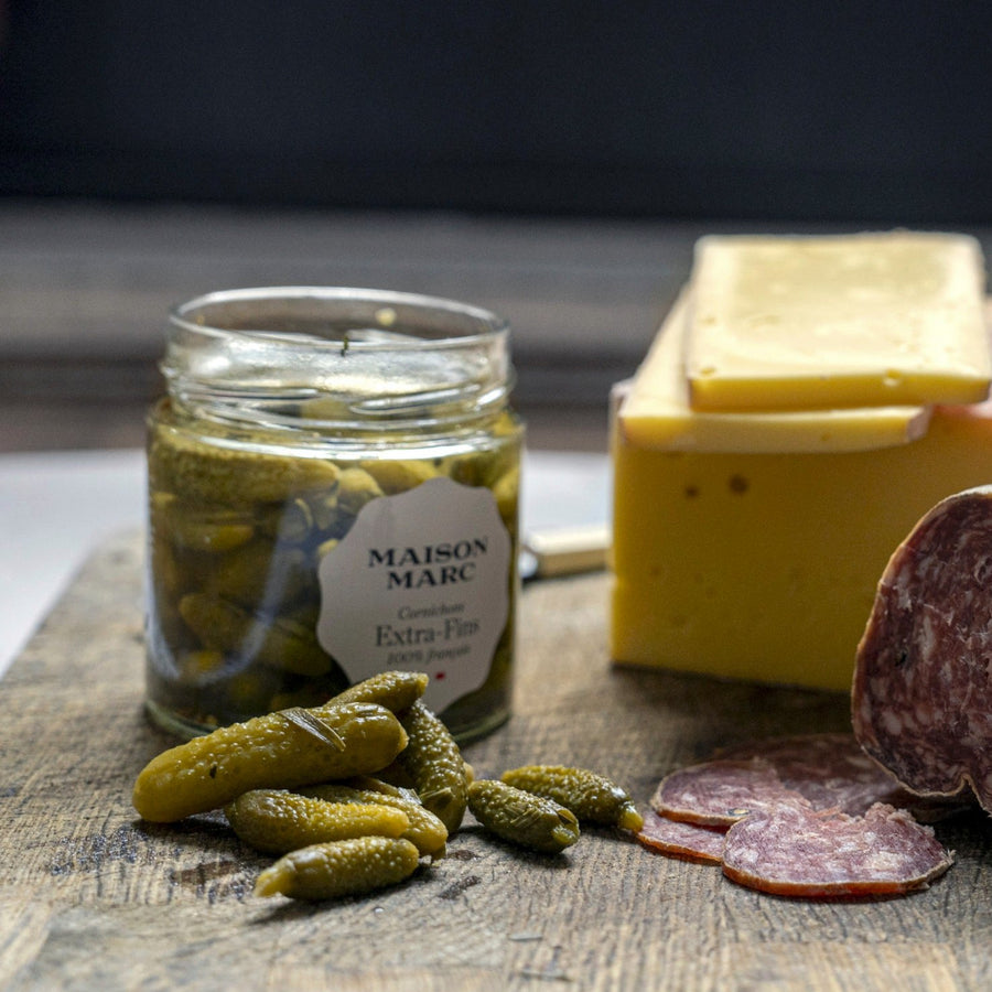 A jar of Maison Marc Extra Fins cornichons on a wooden board with some sliced salami and cheese.