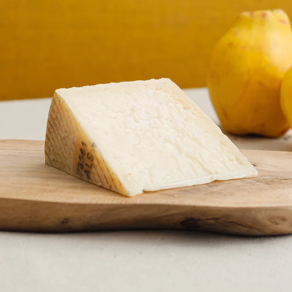 A wedge of creamy off-white cheese on a wooden cutting board, with a blurred yellow fruit in the background.