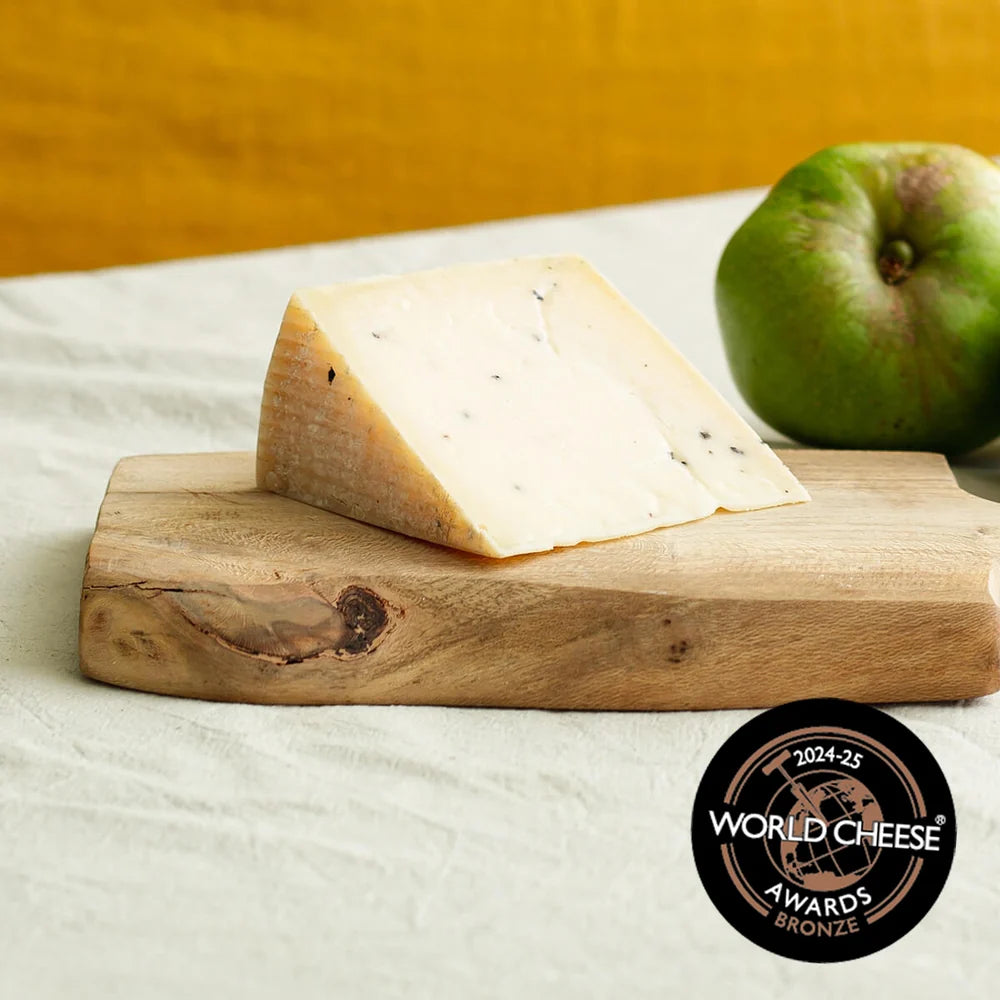 A wedge of blue cheese on a rustic wooden cutting board, with a green apple in the background and a World Cheese Awards 2024-25 Bronze badge in the lower right corner.