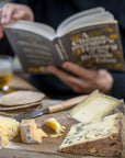 A selection of cuts of cheese and biscuits on a wooden board, with a person reading a book in the background.