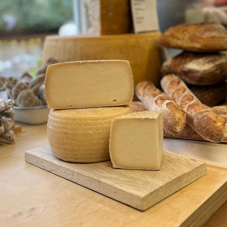A round and rectangular block of pale yellow cheese displayed on a wooden board, with a loaf of rustic bread in the softly blurred background.