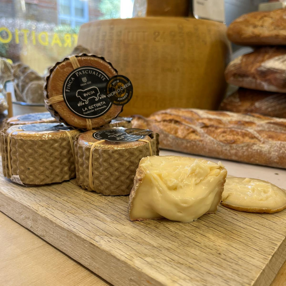 A wooden board displays several small wheels of cheese wrapped in brown paper, with one wheel open to show a pale yellow interior. A round branded label is visible on one wheel, and rustic bread loaves and a large cheese wheel appear blurred in the background inside a bakery or food shop.
