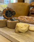 A wooden board displays several small wheels of cheese wrapped in brown paper, with one wheel open to show a pale yellow interior. A round branded label is visible on one wheel, and rustic bread loaves and a large cheese wheel appear blurred in the background inside a bakery or food shop.