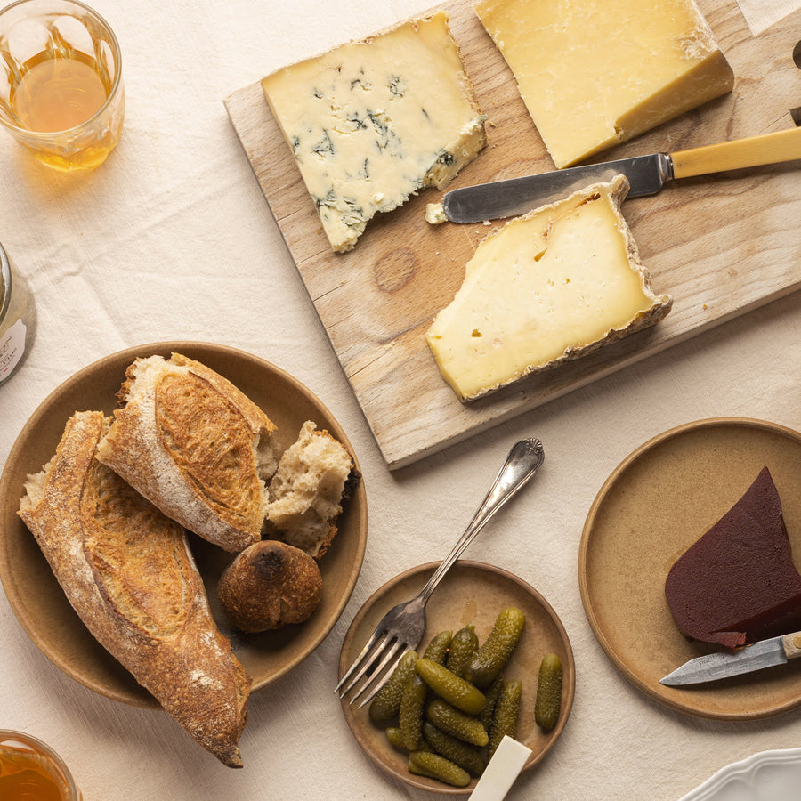 A spread of bread, pickles, quince and cheeses on a wooden board.