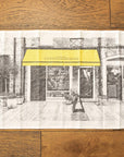 A black and white drawing of a cheese shop printed on a tea towel, placed on a wooden floor.