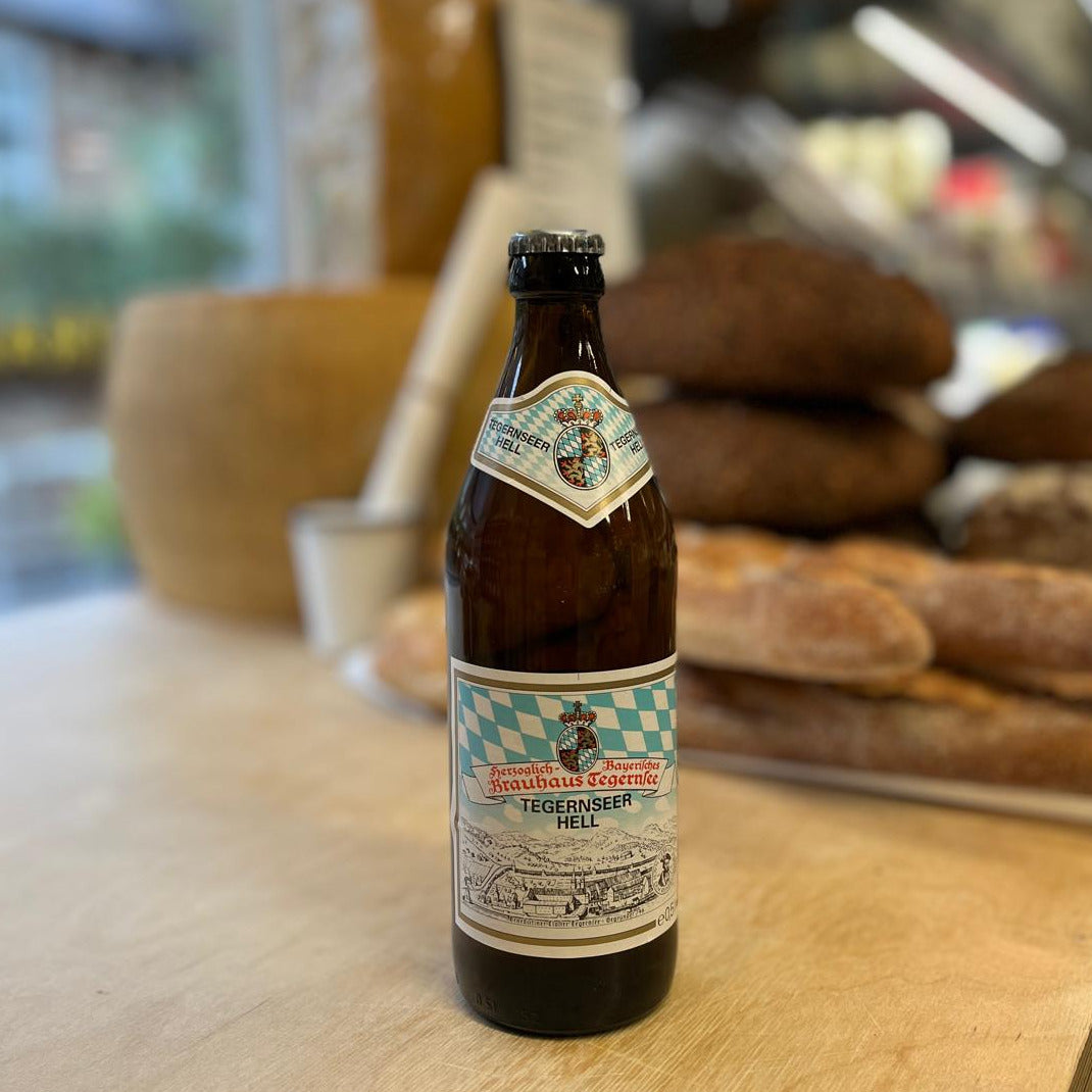 A bottle of Mehlmanns &amp; Söhne Tegernisbeer Melk beer stands on a wooden table with a blurred bakery background featuring loaves of bread.