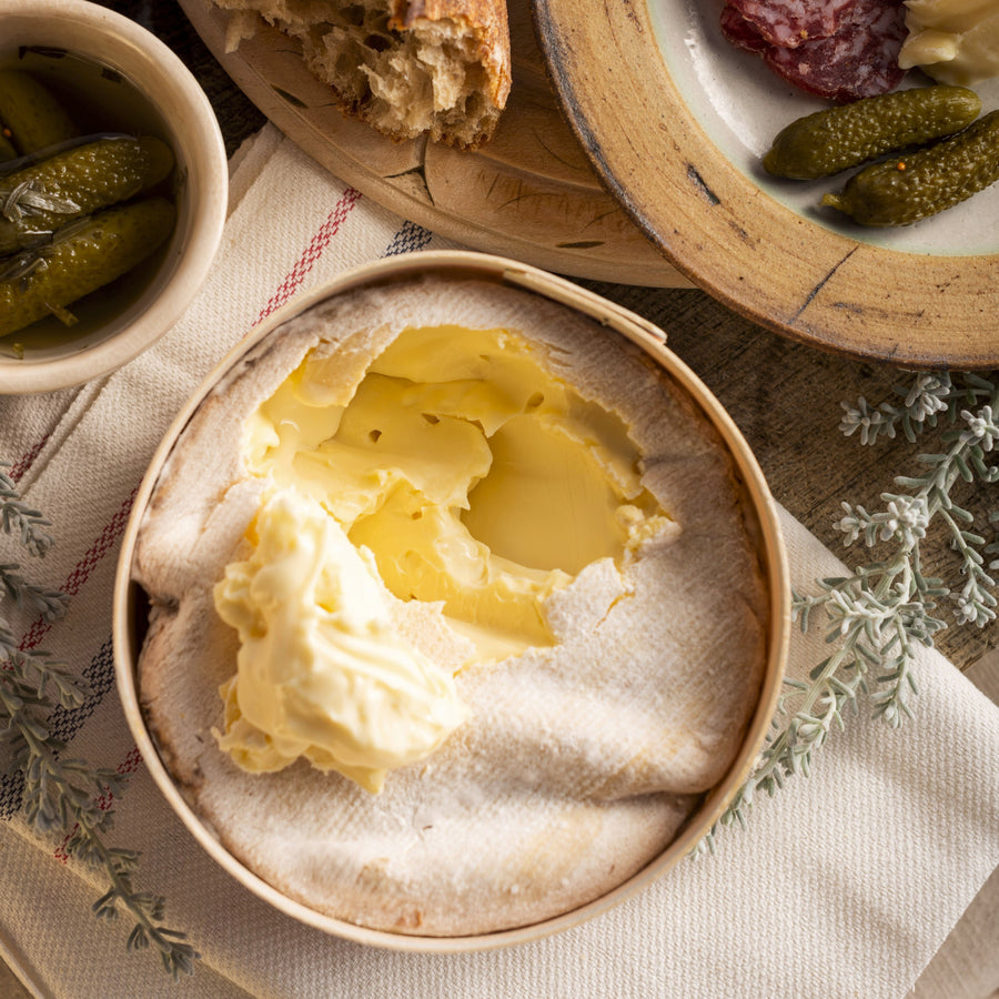 Vacherin cheese with bread, salami, and pickles on a wooden board.