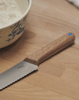 A wooden handled bread knife on a wooden table, next to a patterned ceramic bowl.