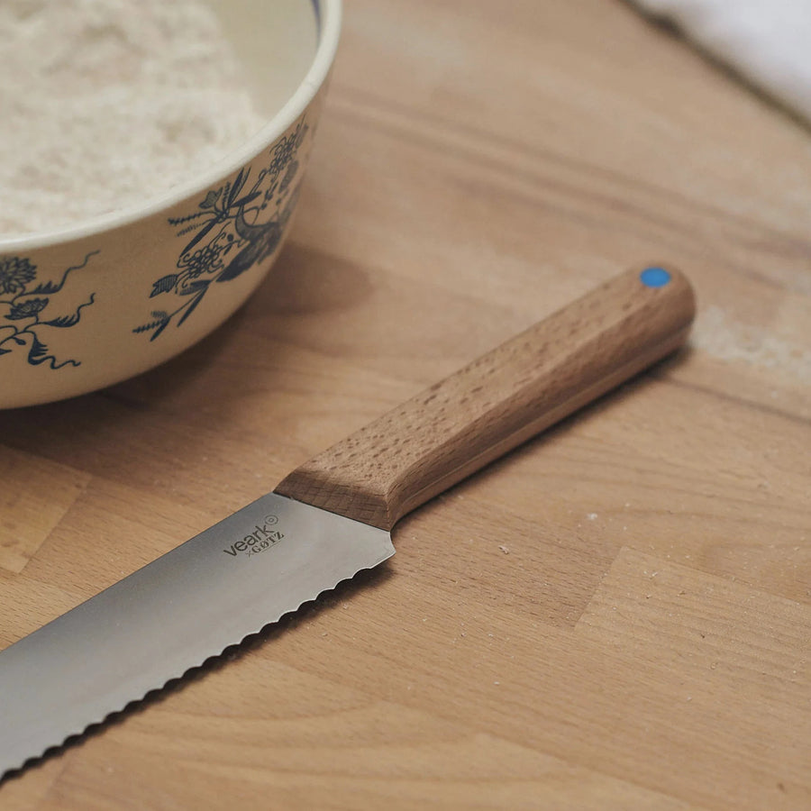 A wooden handled bread knife on a wooden table, next to a patterned ceramic bowl.