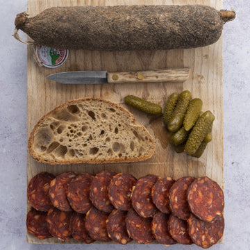 Slices of chorizo on a wooden board with some cornichons, bread and a small knife.