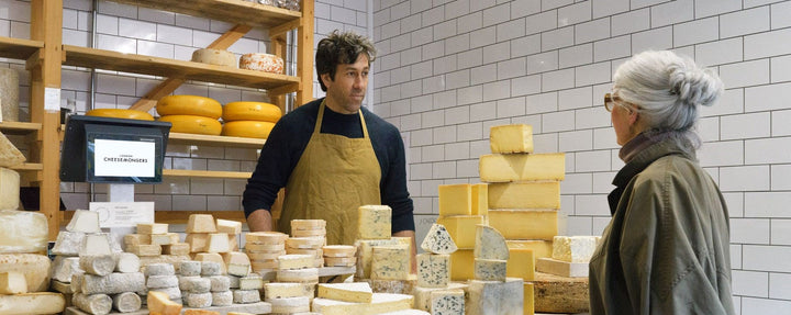 A man standing behind a shop counter of many different cheeses, talking to a grey-haired lady