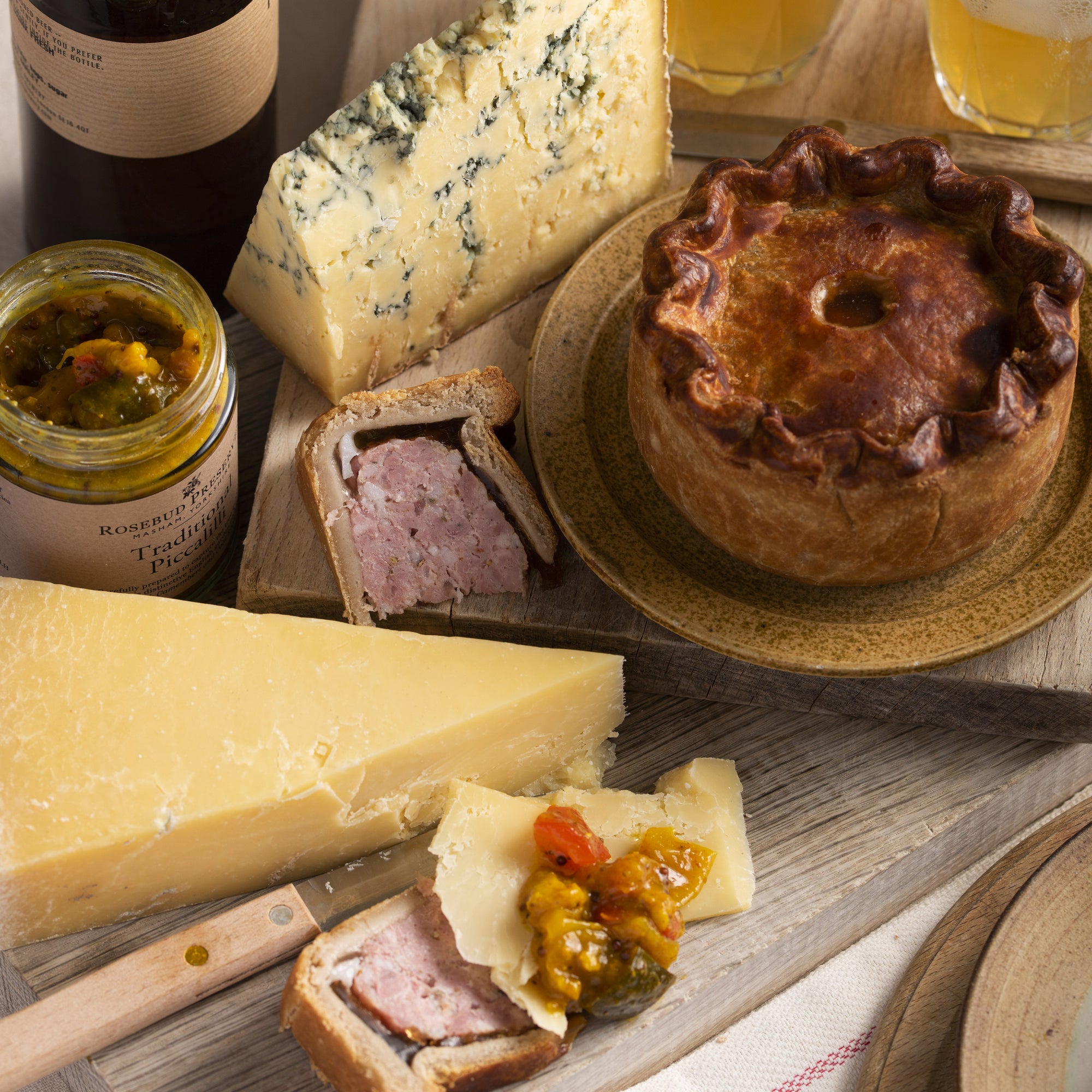 A wooden board laid out with a wedge of Cheddar cheese, a jar of piccalilli, pieces of pork pie and a whole pork pie.