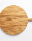 A round wooden chopping board with a small handle at one end on top of a marble worktop.