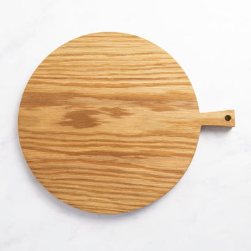A round wooden chopping board with a small handle at one end on top of a marble worktop.