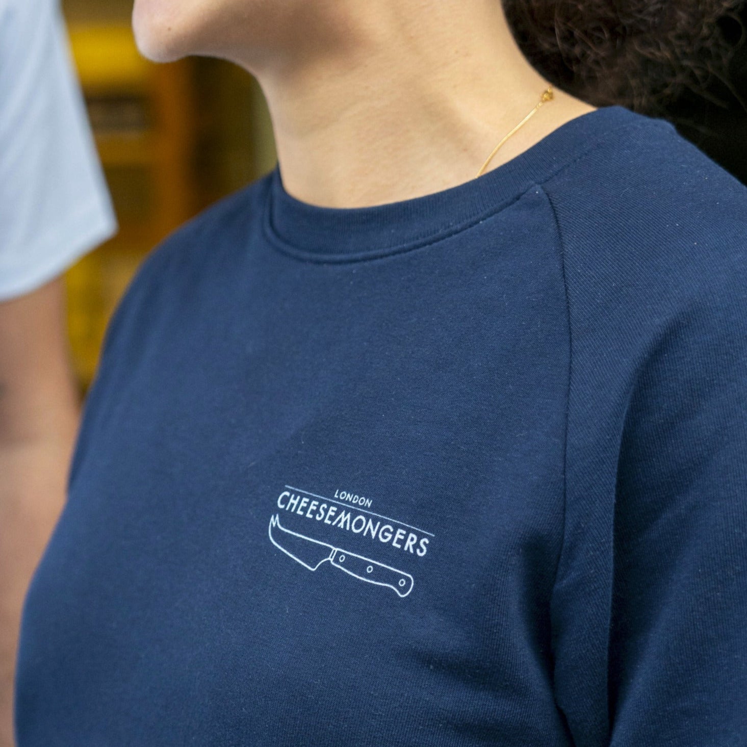 A lady wearing a navy sweatshirt, with a straight London Cheesemongers and knife logo printed on the breast.