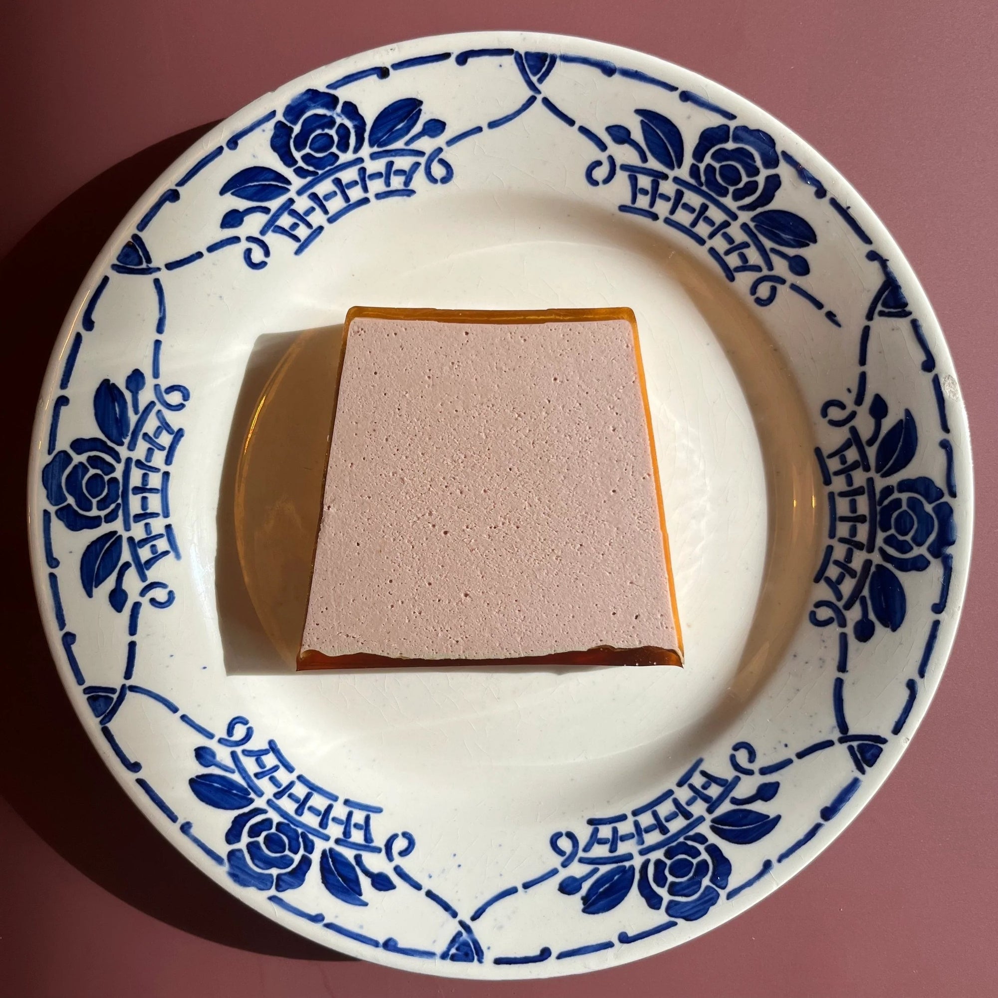 Square piece of duck paté on a white plate with blue floral patterns against a pink background