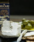 Cuts of Ricotta cheese on biscuits with green olives and a bottle of olive oil in the background.