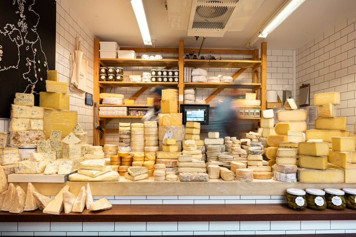 A shop counter containing many different cheeses, with shelves of cheese and jars in the background.