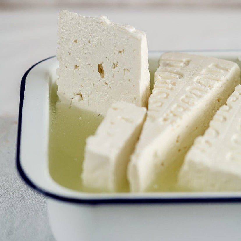 Pieces of Feta cheese in brine in a white enamel dish with a dark blue rim.